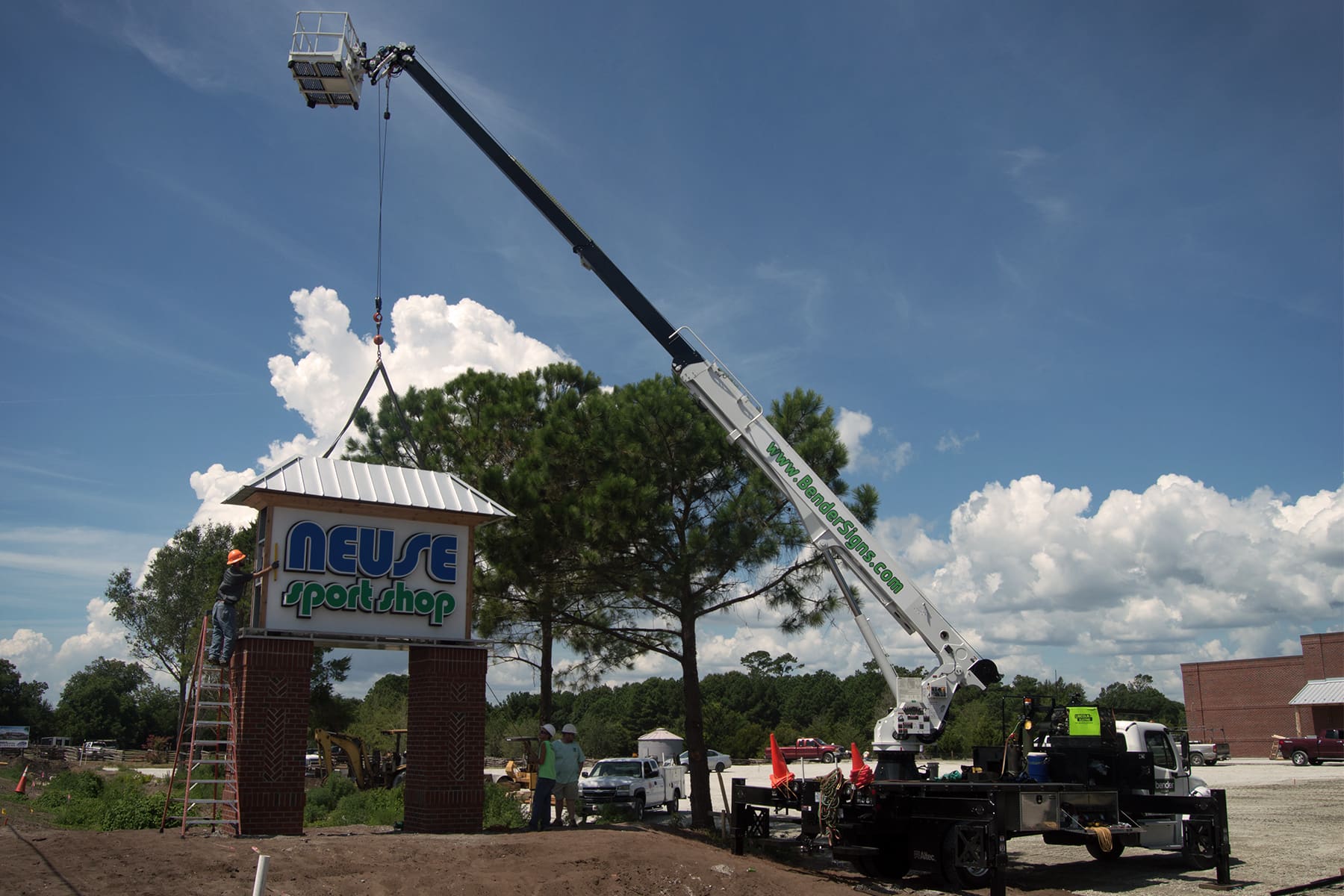 Crane lifting the Neuse Sport Shop pylon cabinet onto brick pillars under storm clouds